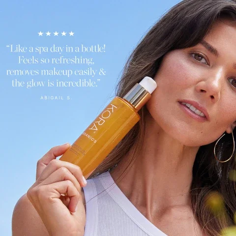 A woman holding a bottle of KORA Organics product stands against a blue background, with the text expressing a positive review that reads, Like a spa day in a bottle! Feels so refreshing, removes makeup easily & the glow is incredible, followed by ABIGAIL S.